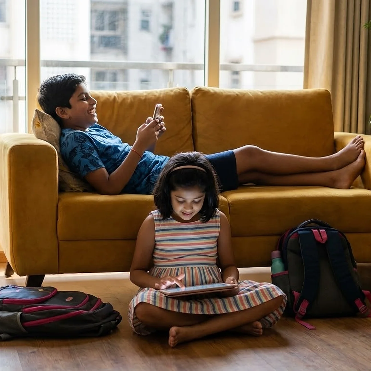 A boy lying on a yellow sofa laughing at a game on his phone while his younger sister sits on the floor with a tablet, their school bags beside her