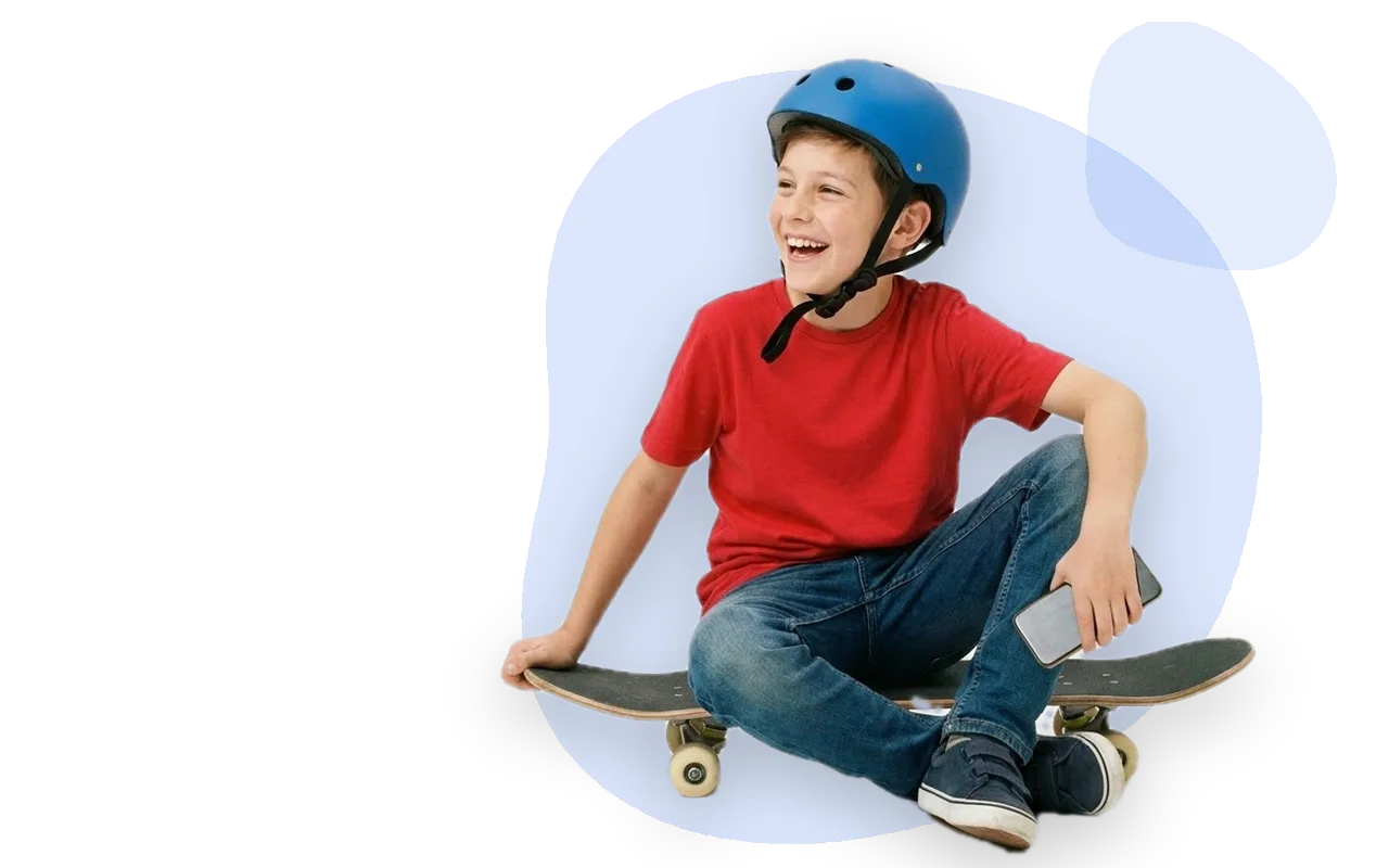 A boy in a blue helmet sitting on his skateboard, phone loose in one hand, laughing at someone out of frame.